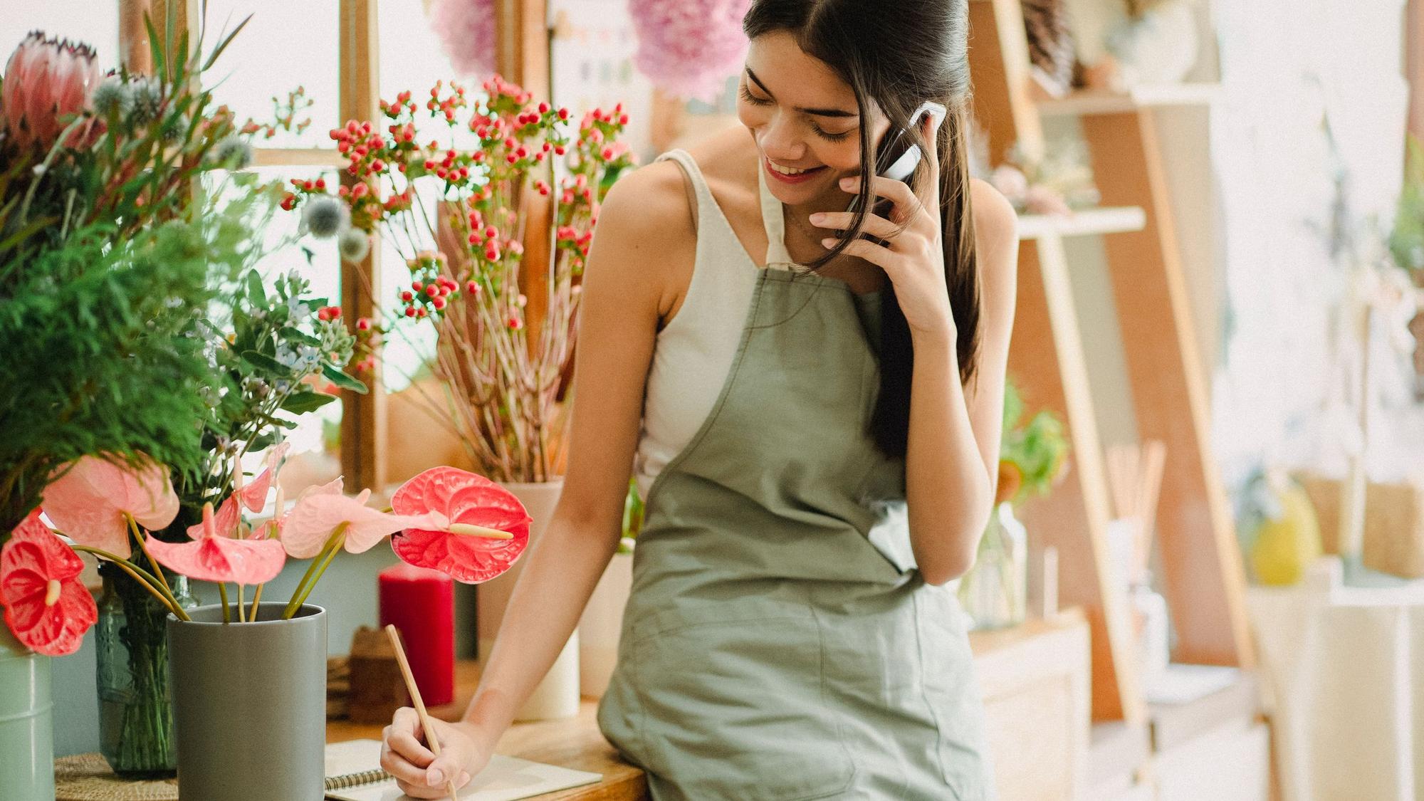 A florist in an apron talks on the phone and takes notes in her shop