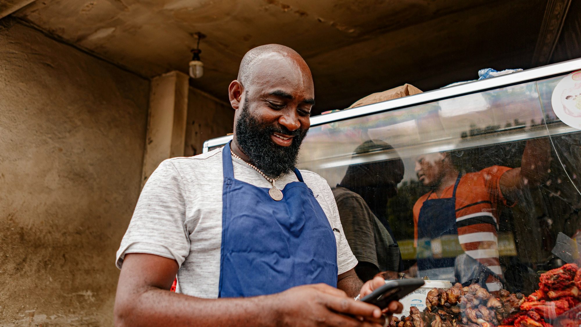 A smiling food vendor in a blue apron checks his phone at his stall