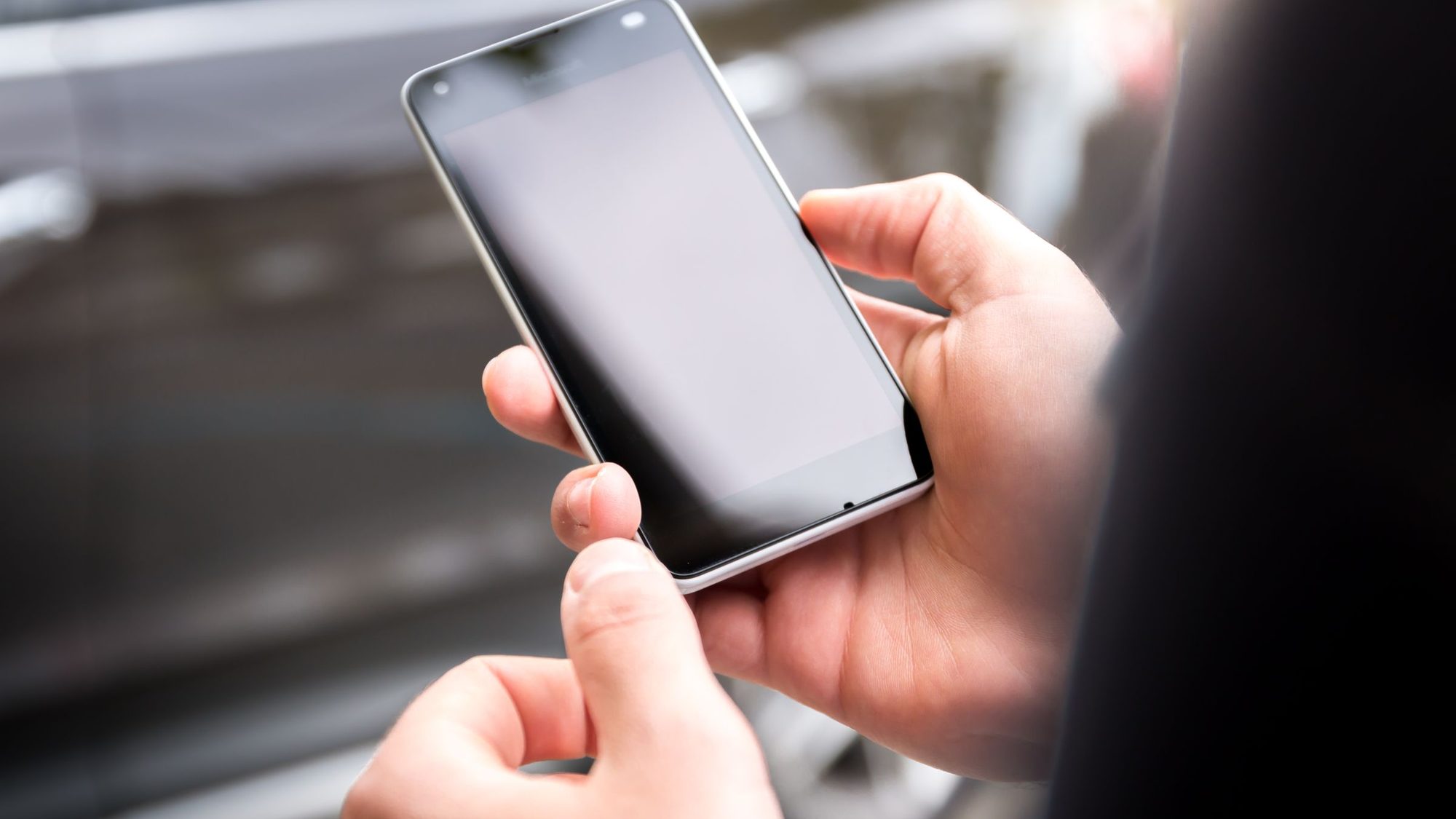 Two hands hold a smartphone with a dark screen outdoors