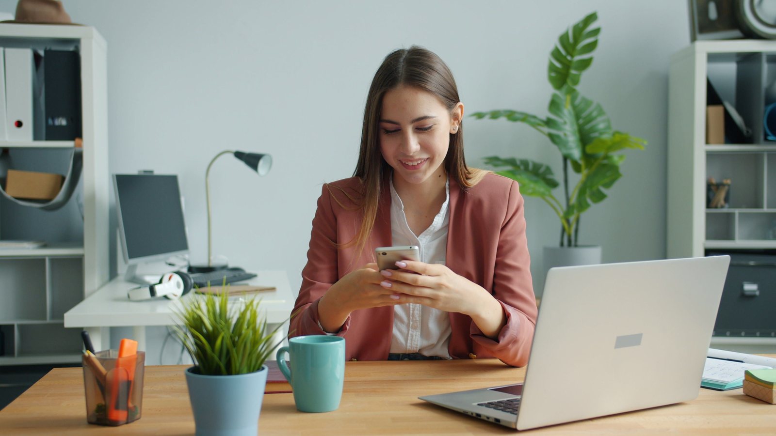 A person at a desk smiling at a smartphone, with a laptop open beside them